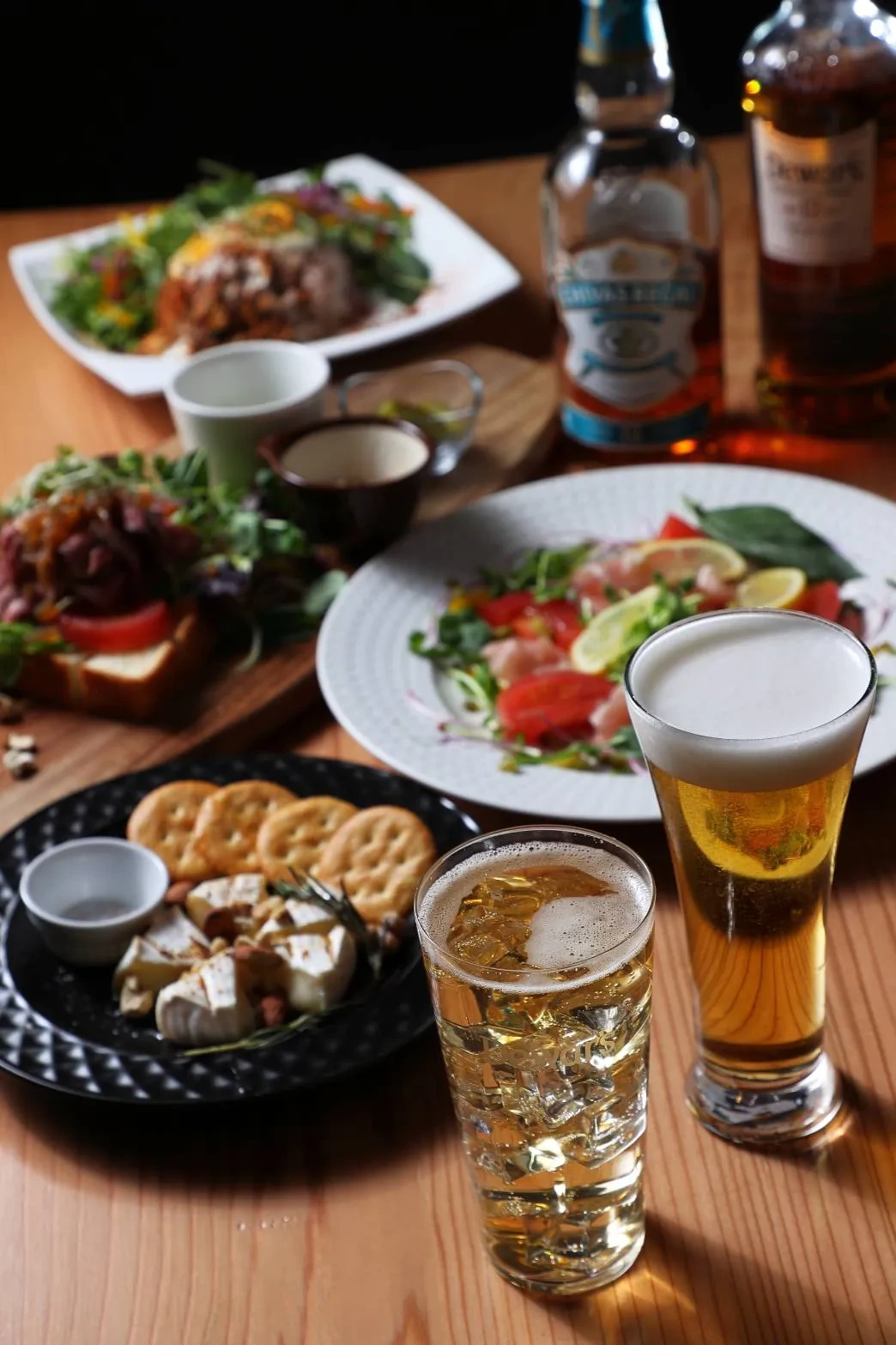 一枚板のテーブルに並んだビール、ハイボール、ウイスキーのボトル、チーズの盛り合わせ、生ハムの一皿、オープンサンド